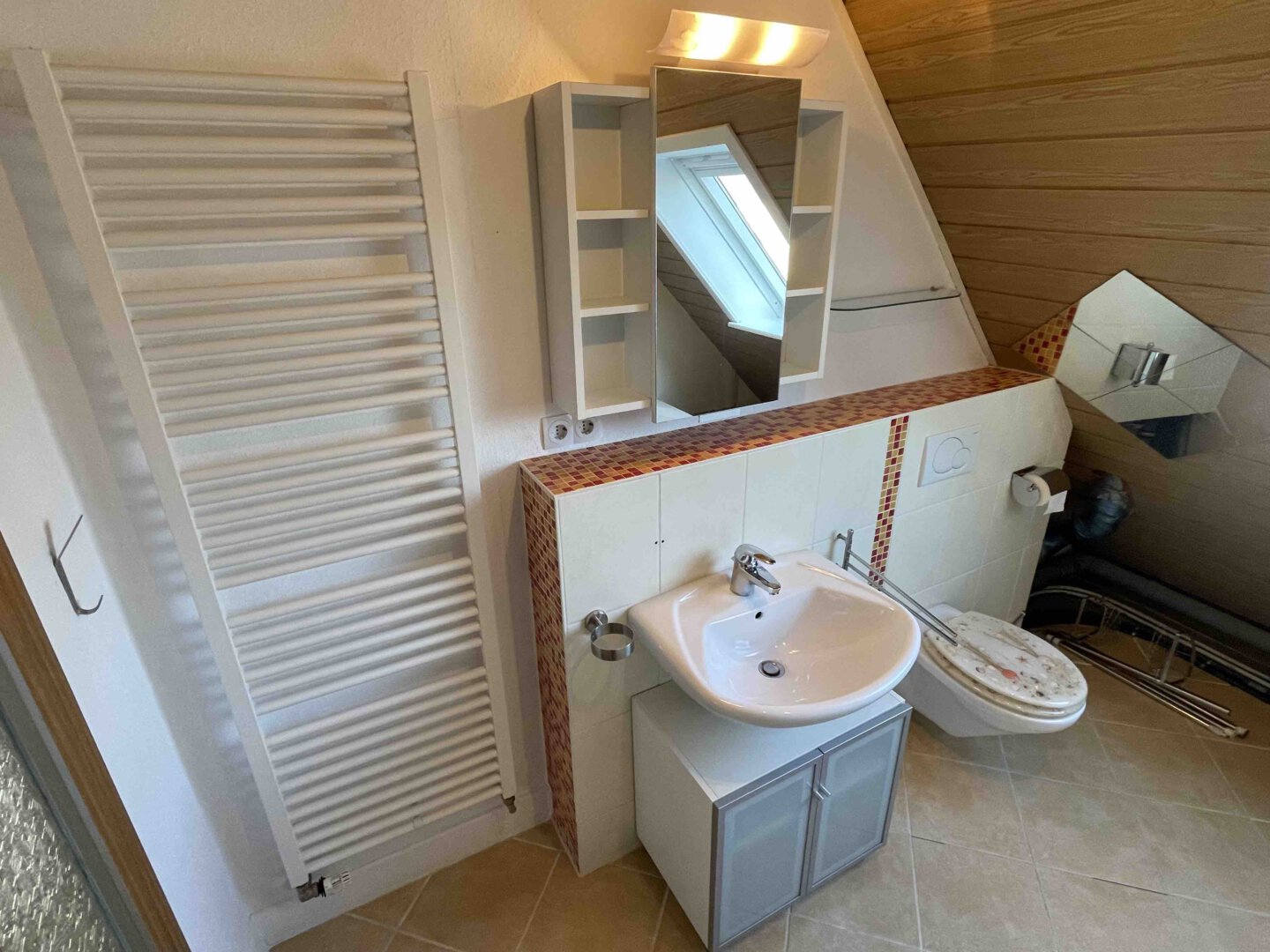 Ein Badezimmer in einem Dreifamilienhaus mit Waschbecken, Spiegel, Toilette, Handtuchheizkörper, Schrank und Dachfenster in der Schräge