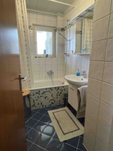 Kleines Bad im Nebengebäude mit blauem Fliesenboden, weißem Waschbecken, Badewanne mit Dusche und Handtuch an der Tür