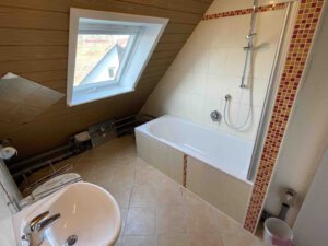 Im Badezimmer eines Dreifamilienhauses mit Dachschräge, Dachfenster, Badewanne mit Dusche, weißem Waschbecken, beigen Fliesen und roten Mosaikakzenten