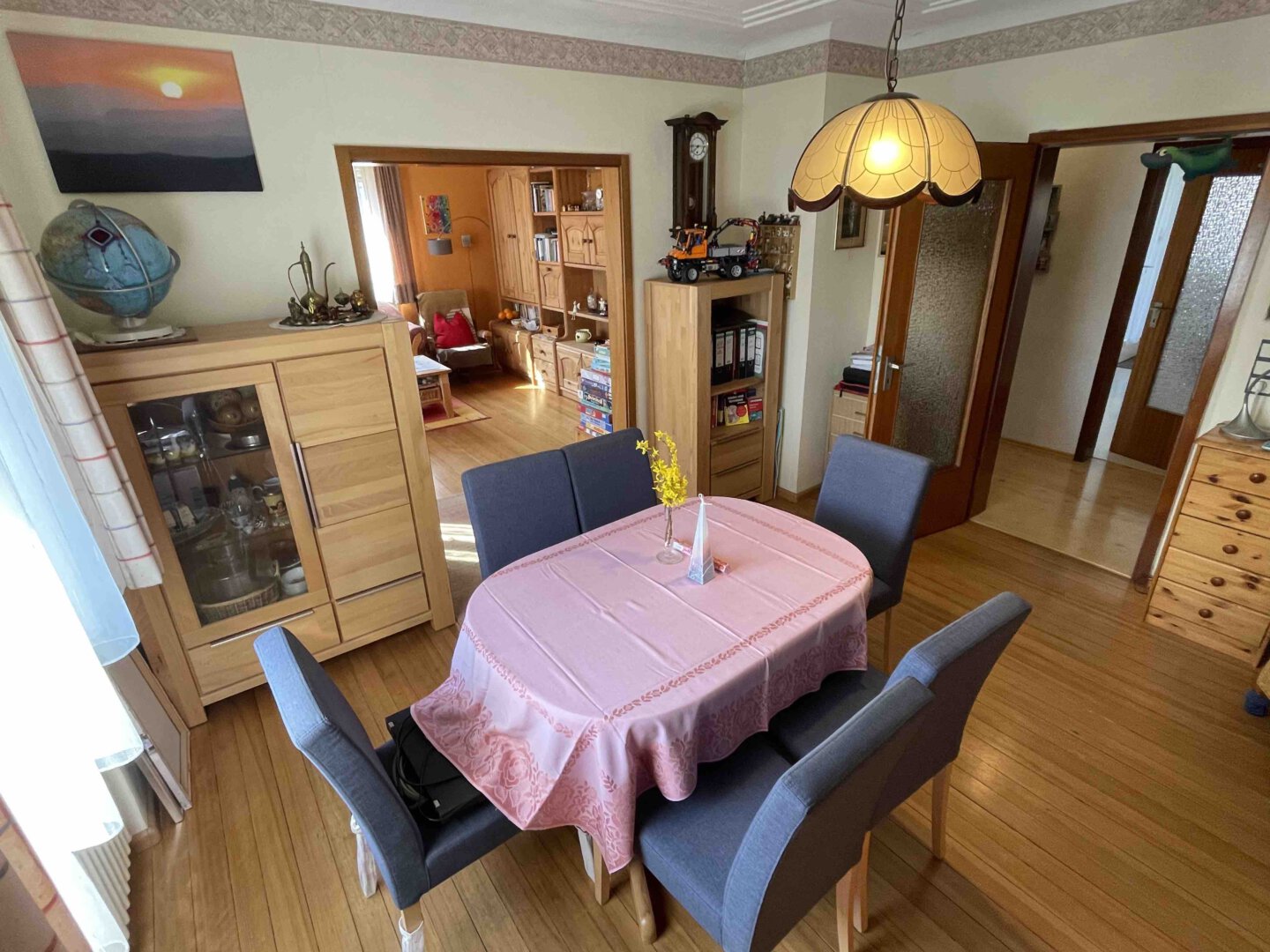 Im Esszimmer eines Dreifamilienhauses liegen ein rosa Tischtuch, graue Stühle, Holzmöbel und eine leuchtende Deckenlampe, dahinter das Wohnzimmer