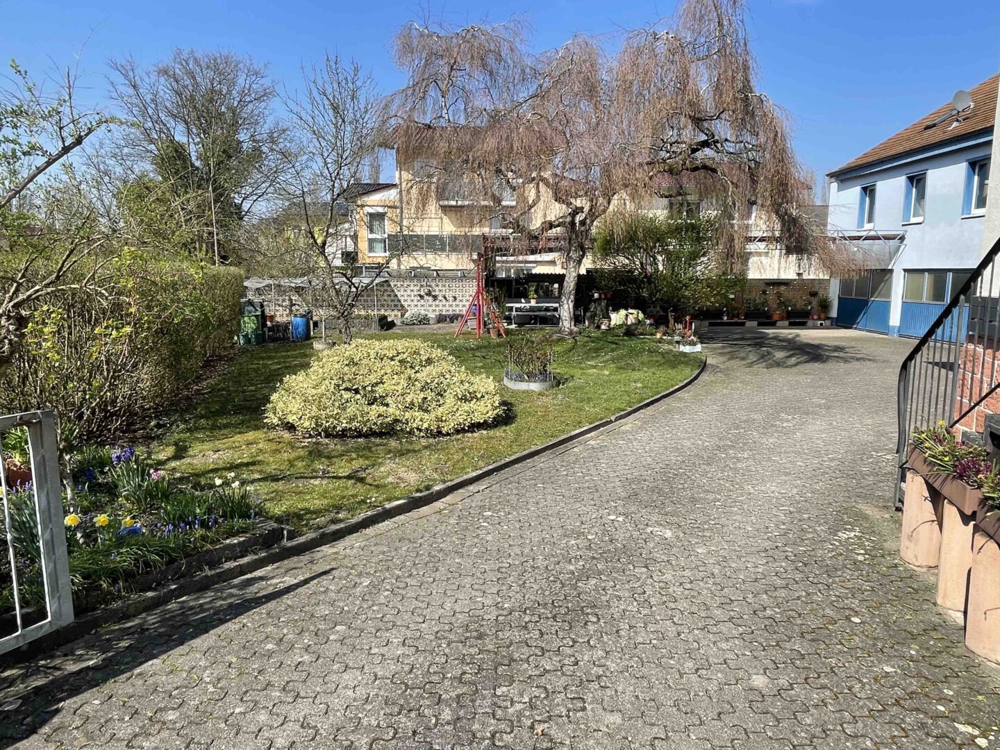 Eine gepflasterte, gebogene Einfahrt mit Blumenbeeten, Bäumen, Garagen und einem blauen Dreifamilienhaus unter klarem Himmel