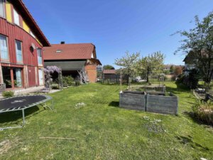 Im sonnigen Garten in Schwanau steht ein Trampolin auf grünem Rasen, blühende Bäume und Doppelhaushälfte-Holzhäuser im Hintergrund