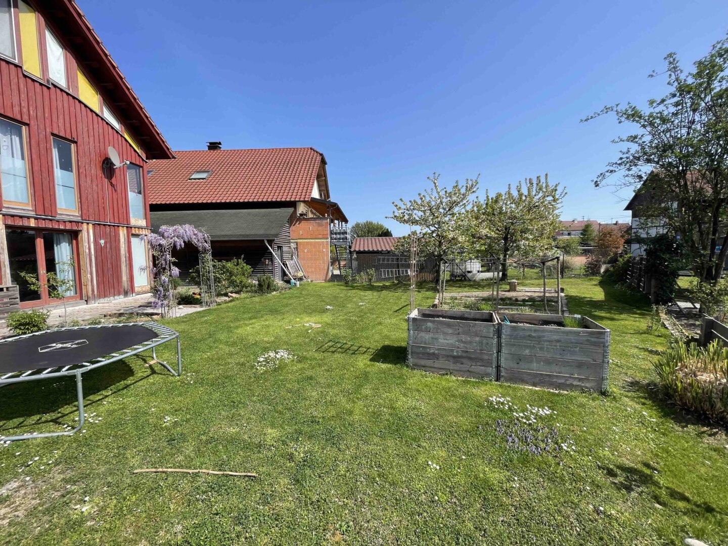 Im sonnigen Garten in Schwanau steht ein Trampolin auf grünem Rasen, blühende Bäume und Doppelhaushälfte-Holzhäuser im Hintergrund