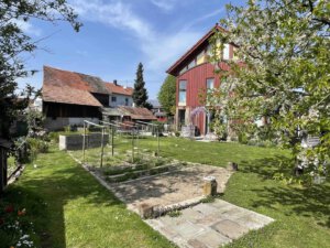 Im sonnigen Garten in Schwanau steht ein rotes Architektenhaus mit großen Fenstern, daneben grüner Rasen und blühender Baum