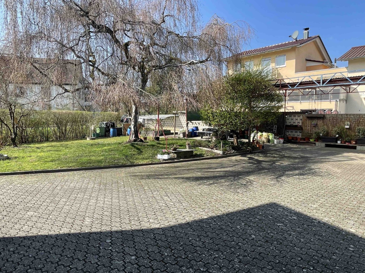 Sonniger Garten mit grünem Rasen, großem Baum, Gartenzubehör sowie einer Garage und einem Dreifamilienhaus im Hintergrund