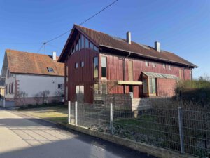 Ein rotes Architektenhaus mit großen Fenstern und schrägem Dach steht an einem sonnigen Tag neben einem kleineren weißen Haus