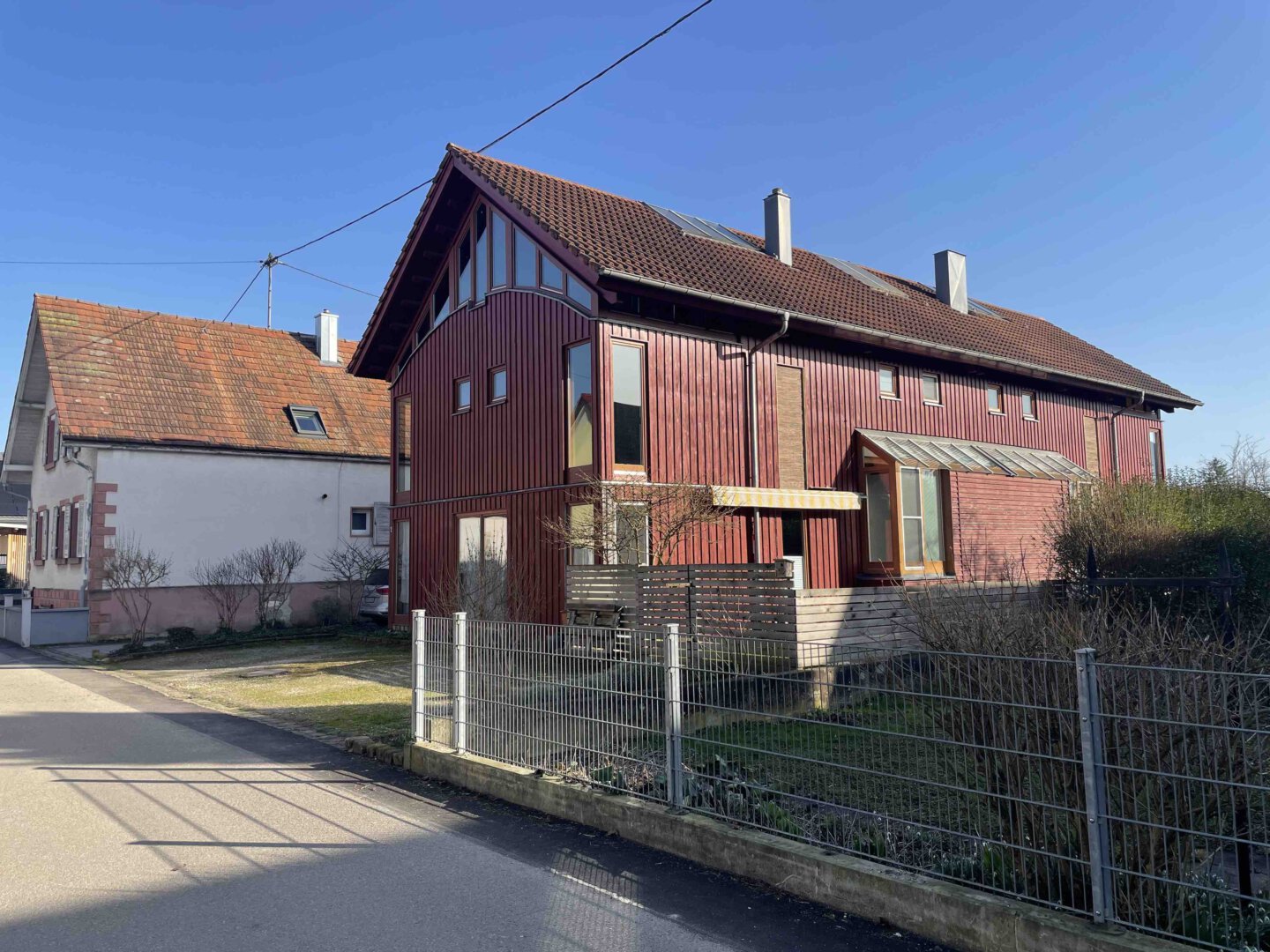 Ein rotes Architektenhaus mit großen Fenstern und schrägem Dach steht an einem sonnigen Tag neben einem kleineren weißen Haus