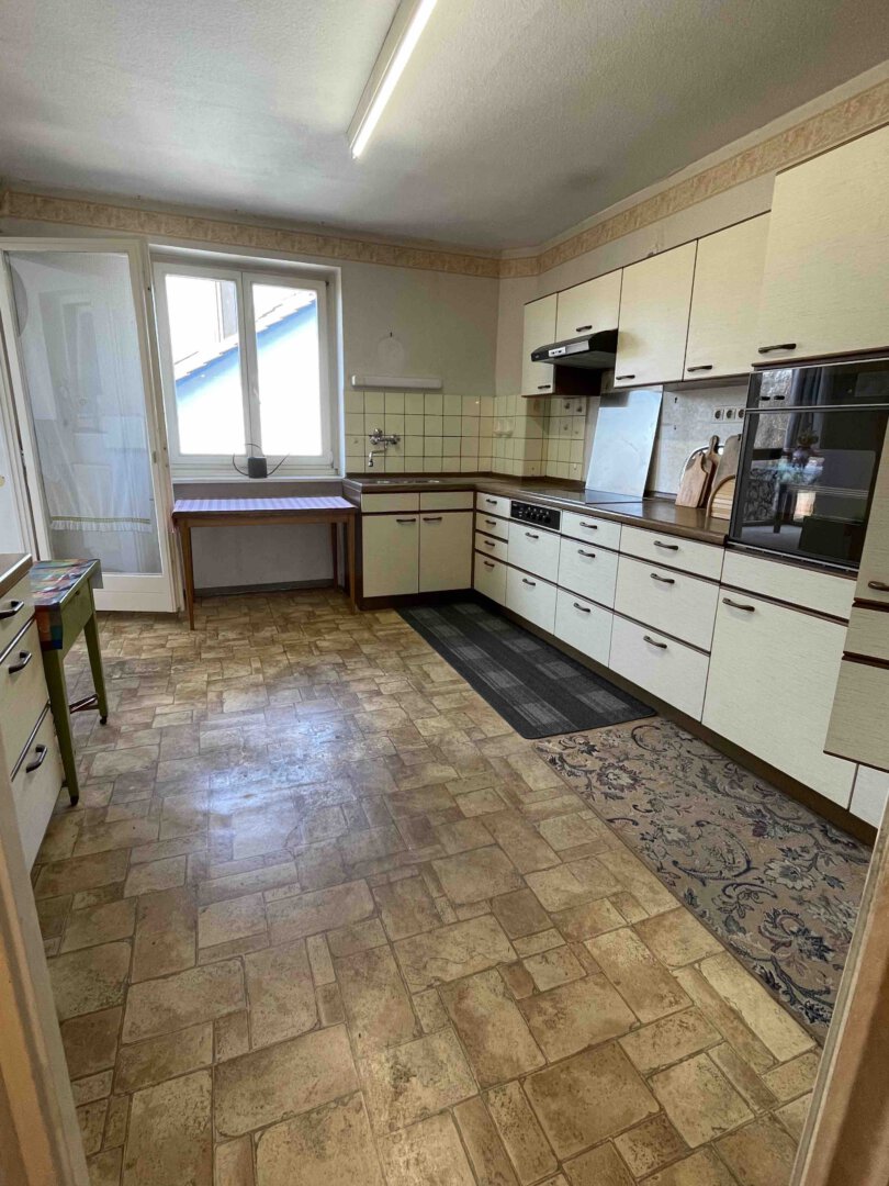 Eine Vintage-Küche in einem Dreifamilienhaus mit cremefarbenen Schränken, gemustertem Linoleum, zwei Teppichen und grünem Tisch an der Tür