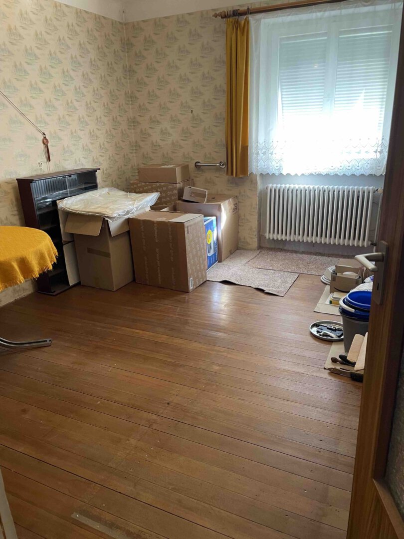 Fast leeres Zimmer mit Holzboden, Umzugskartons und verstreuten Gegenständen in einem Dreifamilienhaus – ideal für Kapitalanleger
