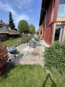 Sonnige Terrasse mit Tisch, Stühlen und Grill an einer roten Doppelhaushälfte, umgeben von grünem Rasen und Bäumen in Schwanau