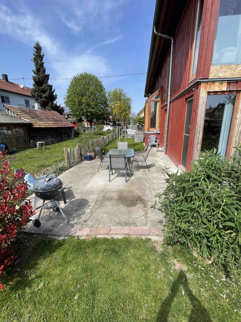 Sonnige Terrasse mit Tisch, Stühlen und Grill an einer roten Doppelhaushälfte, umgeben von grünem Rasen und Bäumen in Schwanau