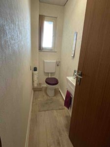 Schmales Badezimmer im Dreifamilienhaus mit Toilette, dunkelviolettem Sitz, kleinem Waschbecken, Fenster, beigen Wänden und Holzboden