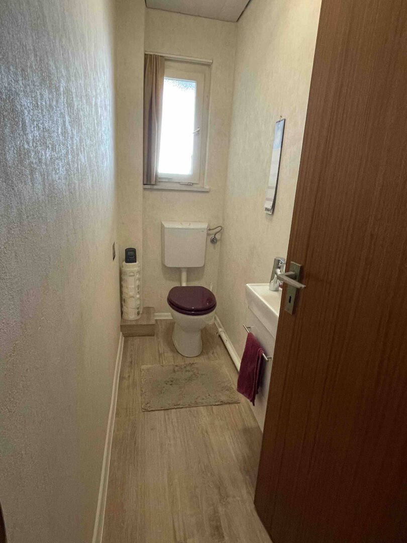 Schmales Badezimmer im Dreifamilienhaus mit Toilette, dunkelviolettem Sitz, kleinem Waschbecken, Fenster, beigen Wänden und Holzboden