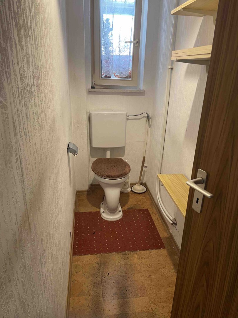 Schmales Bad in einem Dreifamilienhaus mit Toilette, braunem Sitz, rotem Teppich, Regalen rechts und kleinem Fenster oben