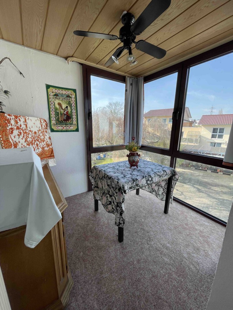 Kleiner Wintergarten in einem Dreifamilienhaus mit großen Fenstern, geblümter Tischdecke, Pflanze, Deckenventilator und Wandbehang