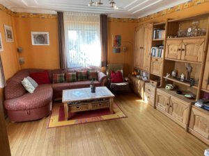 Ein gemütliches Wohnzimmer im Dreifamilienhaus mit orangefarbenen Wänden, Holzmöbeln, rotem Sofa, Bücherregalen und Sonnenlicht