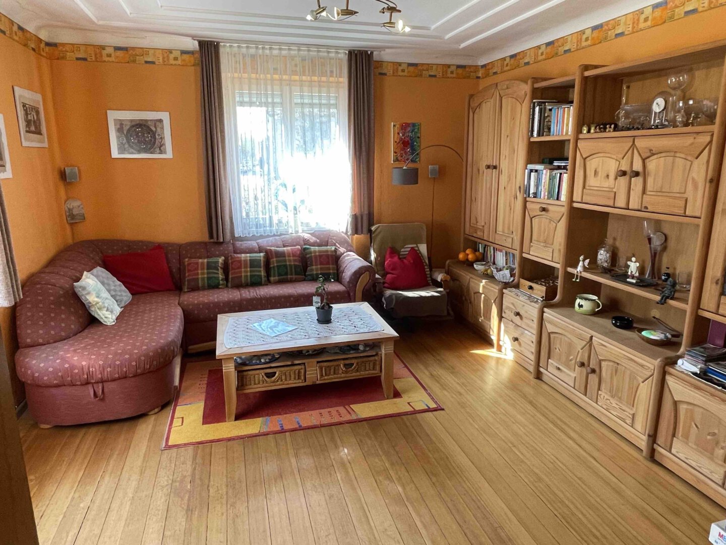 Ein gemütliches Wohnzimmer im Dreifamilienhaus mit orangefarbenen Wänden, Holzmöbeln, rotem Sofa, Bücherregalen und Sonnenlicht