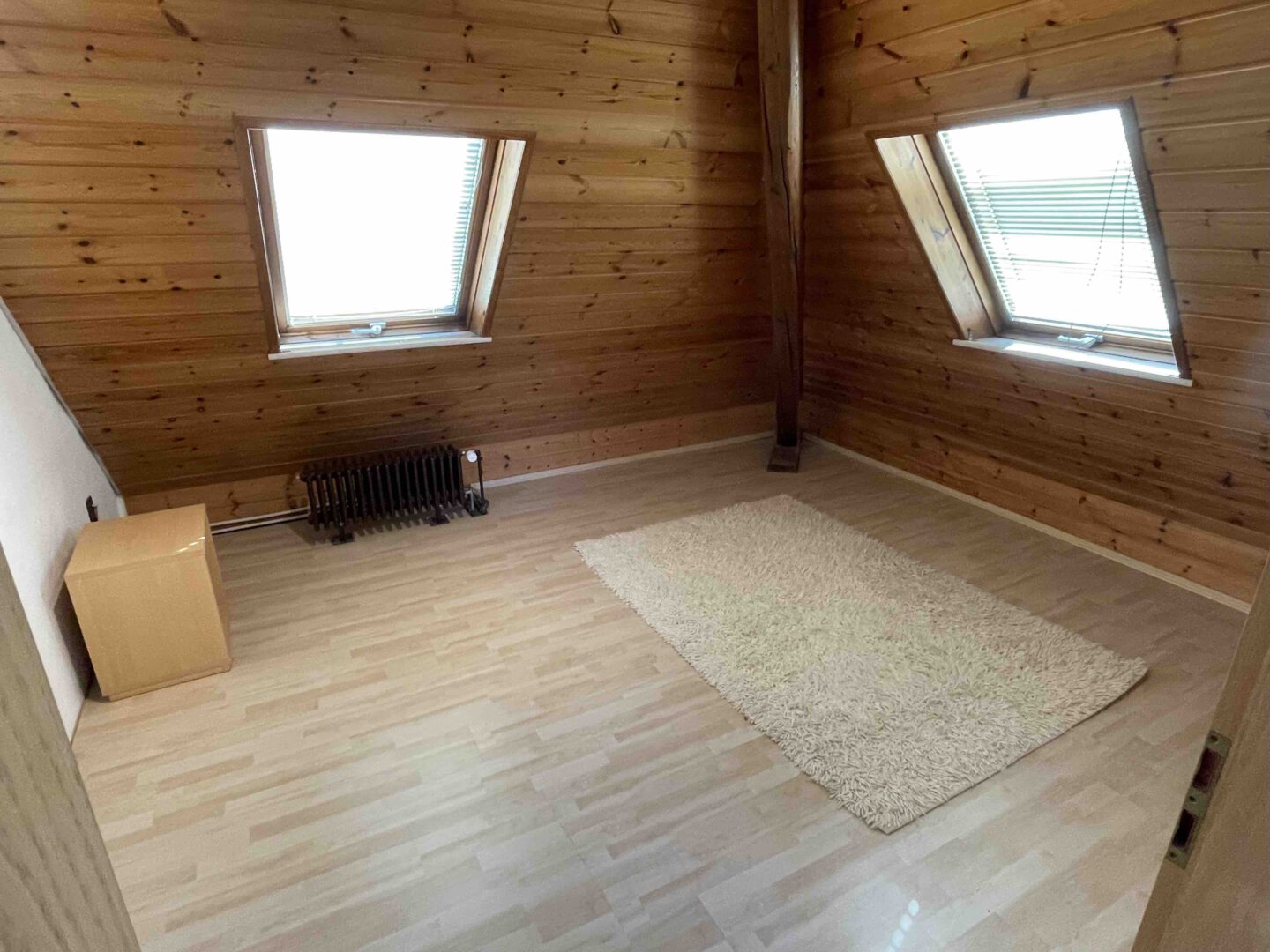 Dachzimmer im Dreifamilienhaus mit holzvertäfelten Wänden, zwei Dachfenstern, Heizung, kleinem Schrank und beigem Teppich auf hellem Holzboden