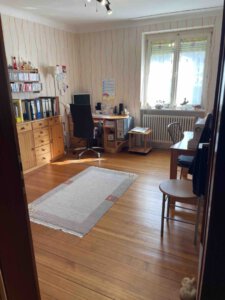 Arbeitszimmer in einem Dreifamilienhaus mit Holzboden, Schreibtisch am Fenster, Regalen und gemütlichem Teppich—ideal für Kapitalanleger