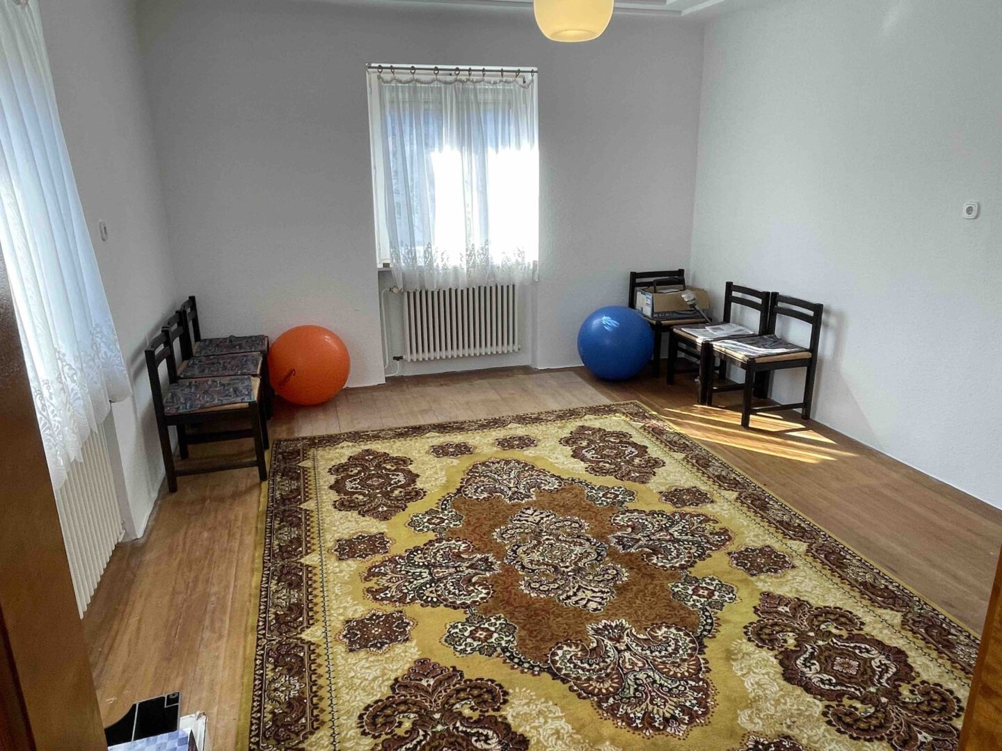 In einem Dreifamilienhaus steht ein spärlich eingerichtetes Zimmer mit gemustertem Teppich, Holzstühlen und lichtdurchfluteten Vorhängen