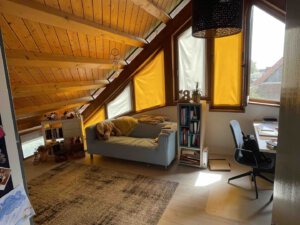 Helles Dachzimmer in einem Schwanau Architektenhaus mit schräger Holzdecke, Sofa, Schreibtisch, Bücherregalen und gelben Vorhängen