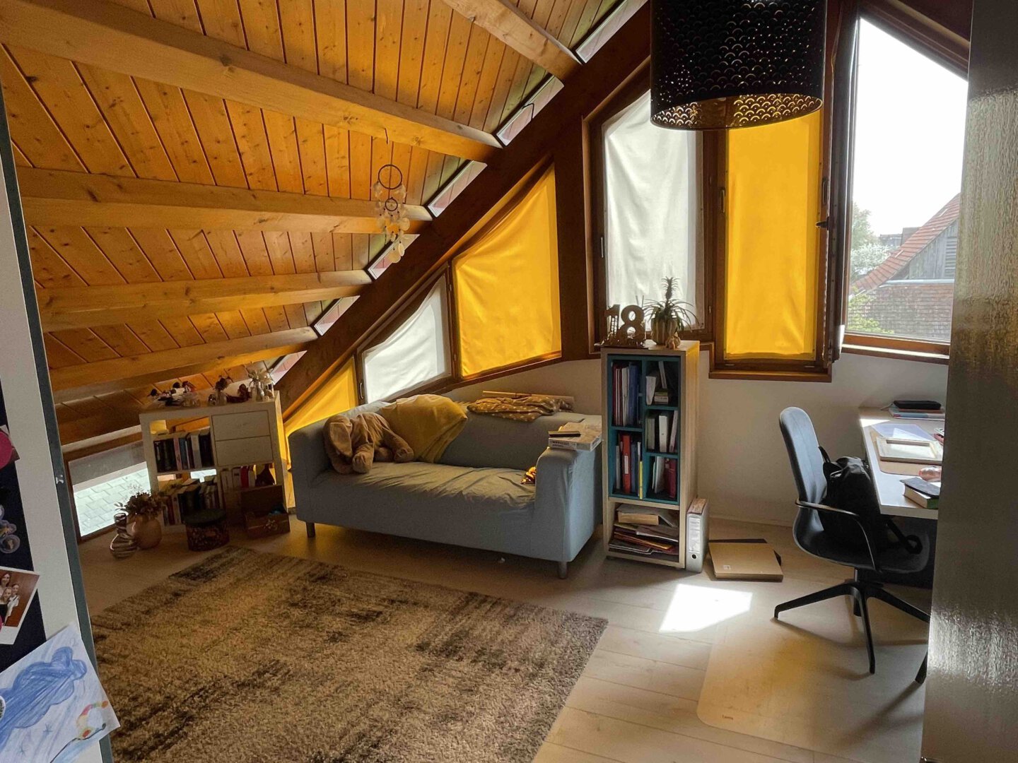 Helles Dachzimmer in einem Schwanau Architektenhaus mit schräger Holzdecke, Sofa, Schreibtisch, Bücherregalen und gelben Vorhängen