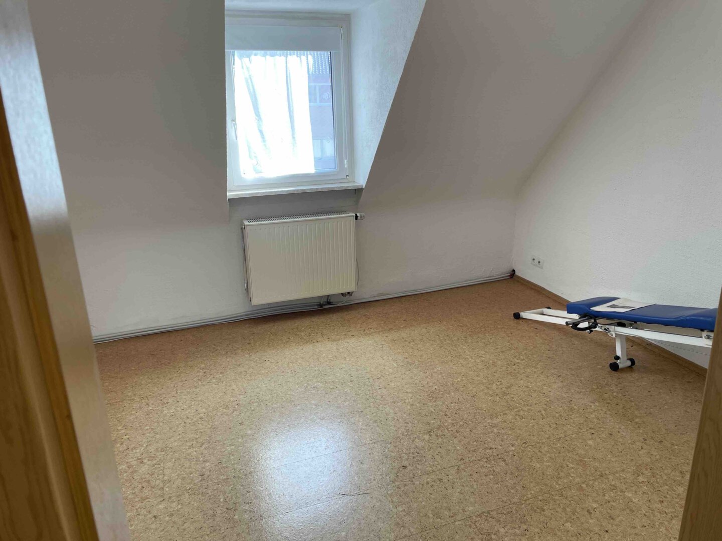 Ein fast leerer Raum in einem Dreifamilienhaus mit Fenster, Heizung und blauer Untersuchungsliege auf braunem Boden