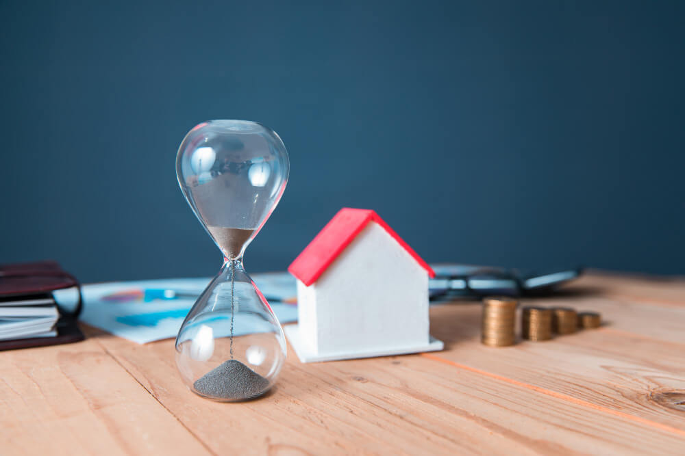Eine Sanduhr, ein kleines Modellhaus, Münzen und Dokumente auf einem Holztisch als automatisch gespeicherter Entwurf