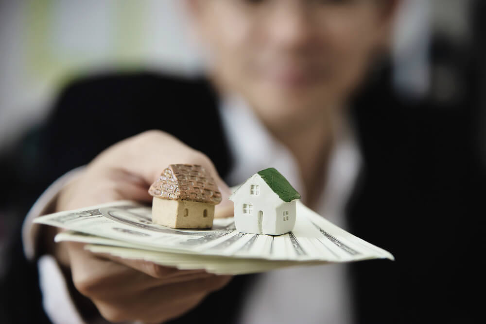 Eine Person hält Modellhäuser und Geldscheine in der Hand, was als Symbol für Immobilieninvestitionen oder den Kauf eines Hauses dient