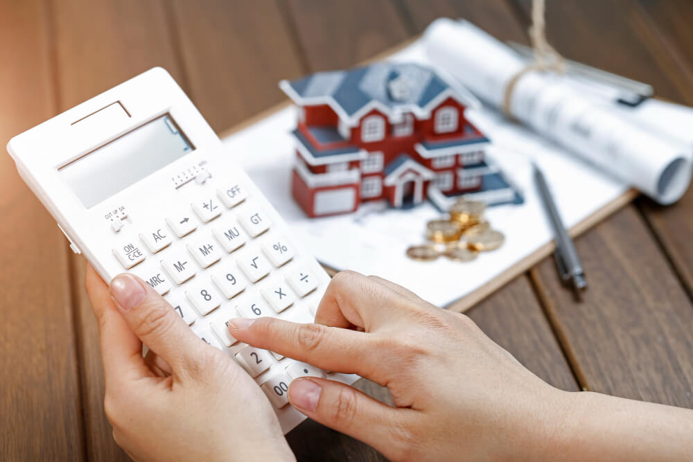 Jemand rechnet mit einem Taschenrechner neben einem Modellhaus, Münzen und Bauplänen – automatisch gespeicherter Entwurf für Ihr Finanzkonzept